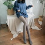 Mannequin brune appuyée sur une table avec une nappe blanche et une plante verte à gauche, portant une robe pull bleu foncé et une paire de collant polaire gris effet transparent. Elle a une jambe tendue, l'autre plié. Une main remonte un peu sa robe. Le sol est en parquet.