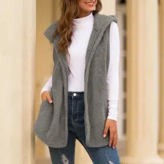 Mannequin femme debout devant un monument de couleur beige, portant un jean foncé, un col roulé blanc, un long gilet gris avec des poches et une capuche, des lunettes de soleil.