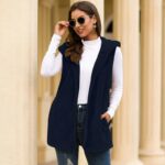 Mannequin femme debout devant un monument de couleur beige, portant un jean foncé, un col roulé blanc, un long gilet avec des poches et une capuche, des lunettes de soleil.