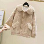 Manteau polaire femme beige, avec col rabattu et poches plaquées avec rabats. Sur un cintre, avec un mur beige en fond et une main de femme qui tient le bout de la manche à gauche.