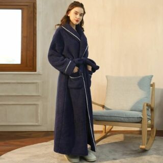 Mannequin femme, debout devant un rockingchair moderne, avec un mur beige dans le fond. Elle porte un long peignoir bleu marine et des chaussons clairs, elle a les mains dans les poches et est de trois-quart.