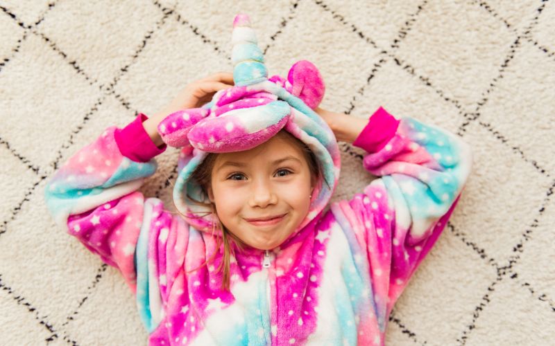 Petite fille allongée sur un tapis, portant un pyjama combinaison licorne, avec capuche, rose, bleu et blanc.